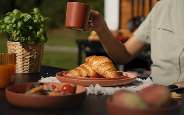 Zwei Croissants auf einem Teller auf einem Gartentisch; daneben ein Glas Orangensaft, eine Schale mit Cherrytomaten und Salat, ein Basilikum in einem geflochtenen Topf und eine Person in hellgrünem T‑Shirt, die eine Tasse hält