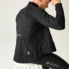 Persona vista da dietro, seduta di lato su uno sgabello bianco, indossa una giacca sportiva nera e leggings neri con loghi bianchi sulla schiena e sulla coscia