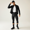Uomo in abbigliamento da ciclismo nero con berretto nero, giacca nera, T‑shirt bianca, pantaloncini da ciclismo neri a tre quarti, calzini e scarpe nere, su sfondo chiaro, si aggiusta il berretto con la mano destra