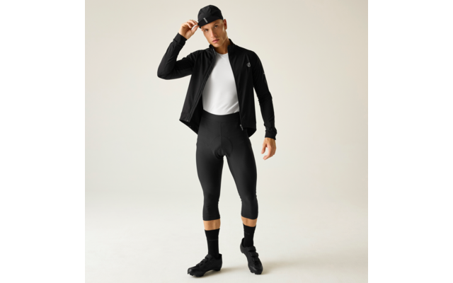 Uomo in abbigliamento da ciclismo nero con berretto nero, giacca nera, T‑shirt bianca, pantaloncini da ciclismo neri a tre quarti, calzini e scarpe nere, su sfondo chiaro, si aggiusta il berretto con la mano destra