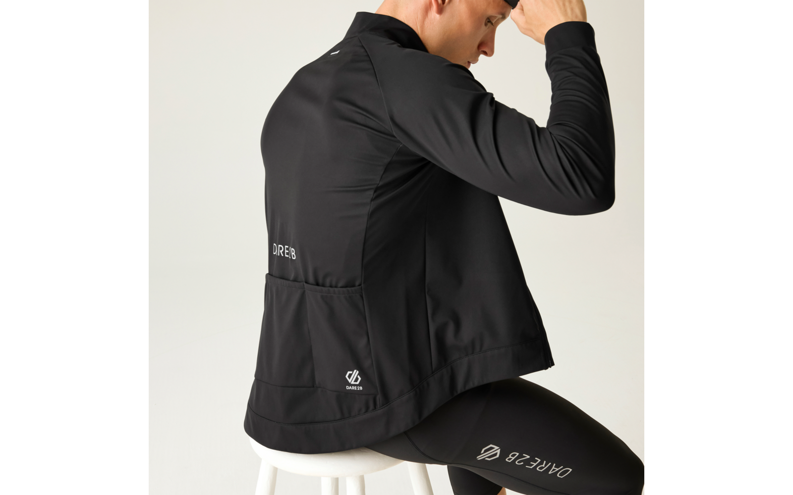 Persona vista da dietro, seduta di lato su uno sgabello bianco, indossa una giacca sportiva nera e leggings neri con loghi bianchi sulla schiena e sulla coscia