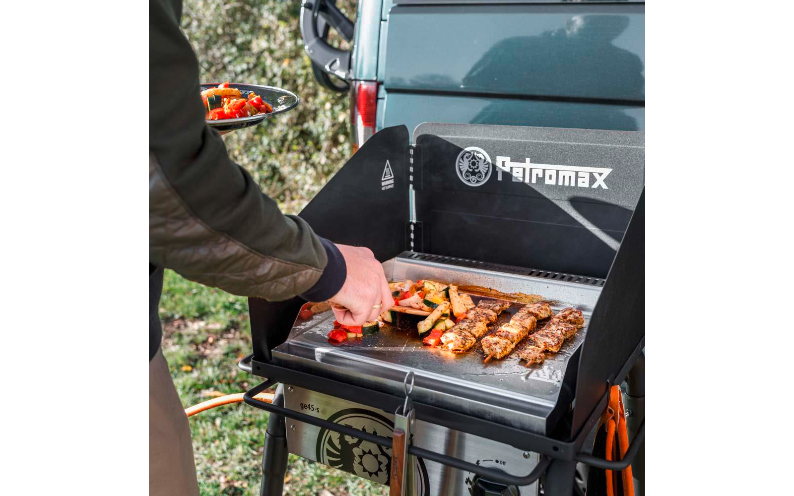 Hand wendet Gemüse und Fleischspieße auf einer Grillplatte neben einem Auto