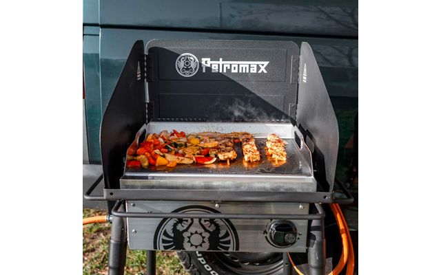 Schwarzer Petromax-Gasgrill mit Metall-Grillplatte; auf der Platte liegen Fleischspieße, geschnittenes Gemüse und Kartoffelscheiben, es steigt Rauch auf