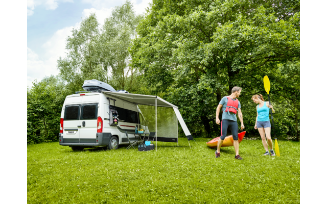 Weißer Campervan mit ausgeklapptem Sonnendach auf einer Wiese; zwei Personen stehen daneben und halten Paddel; zwei bunte Kajaks liegen auf dem Gras; Laubbäume und blauer Himmel im Hintergrund