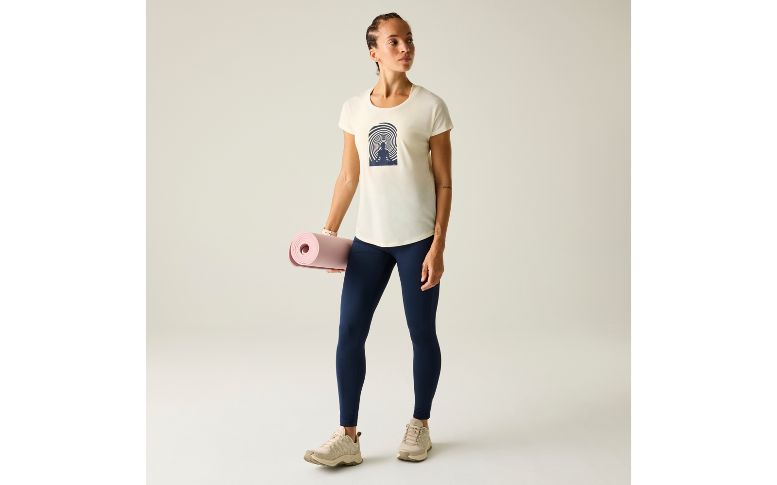 Person steht vor hellem Hintergrund, hält eine gerollte rosa Matte in der linken Hand, trägt cremefarbenes T-Shirt mit dunkelblauem Halbkreis-Motiv, dunkelblaue Leggings und beige Sneaker, Blick nach rechts