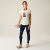 Person steht vor hellem Hintergrund, hält eine gerollte rosa Matte in der linken Hand, trägt cremefarbenes T-Shirt mit dunkelblauem Halbkreis-Motiv, dunkelblaue Leggings und beige Sneaker, Blick nach rechts