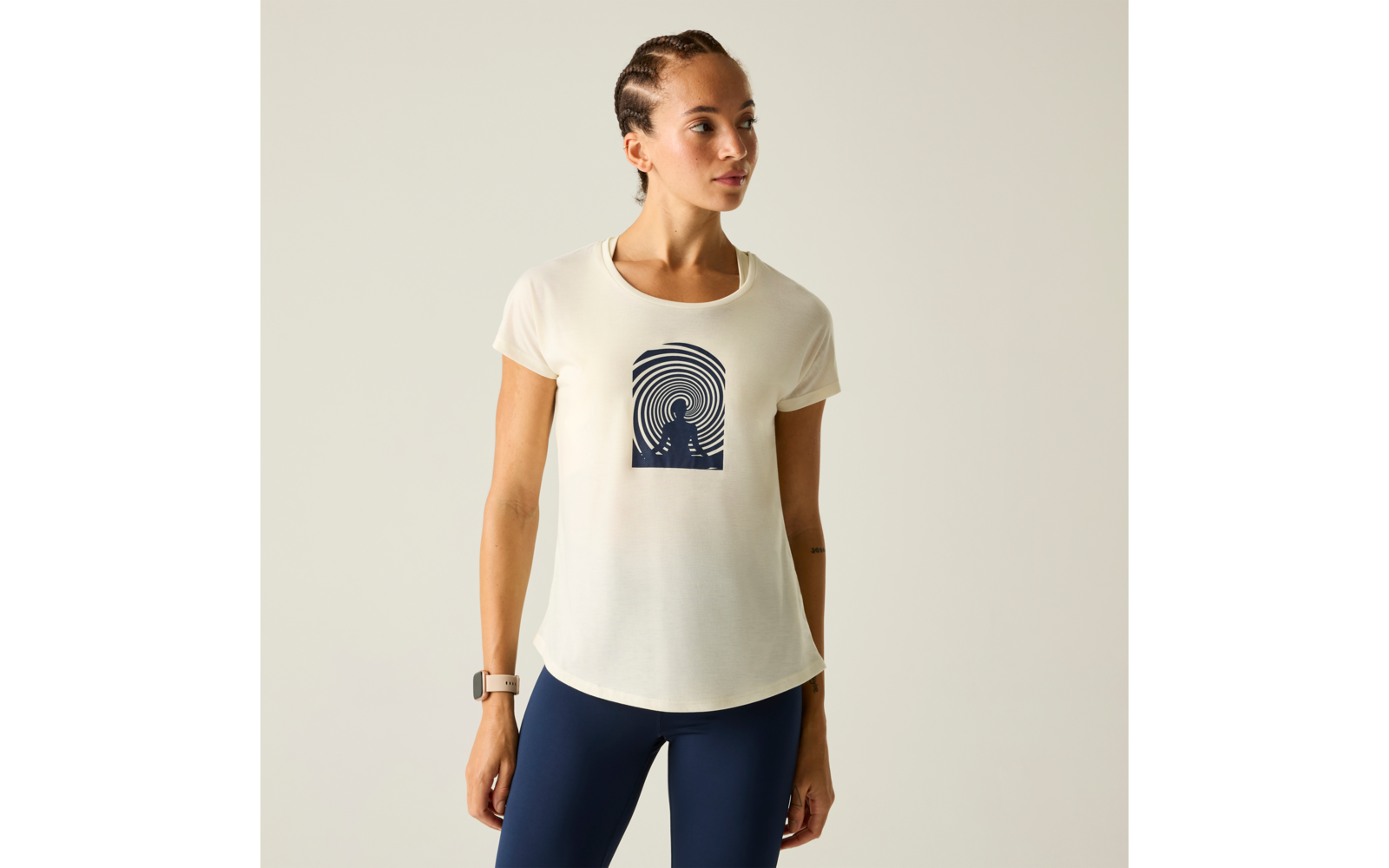 Person mit geflochtenem Haar trägt off-white T-Shirt mit dunklem konzentrischem Kreisdesign und dunkelblauen Leggings vor neutralem Hintergrund