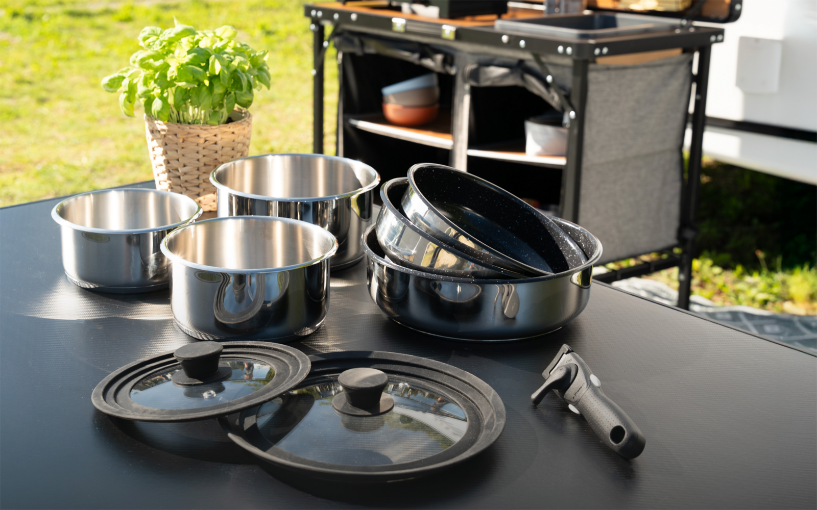 Mehrere Edelstahltöpfe, ineinander gestapelte Pfannen, zwei Glasdeckel mit schwarzen Griffen, ein abnehmbarer Pfannengriff, ein Basilikum in einem Korb auf einem schwarzen Tisch; im Hintergrund ein Küchenwagen mit Regalen und Rasen