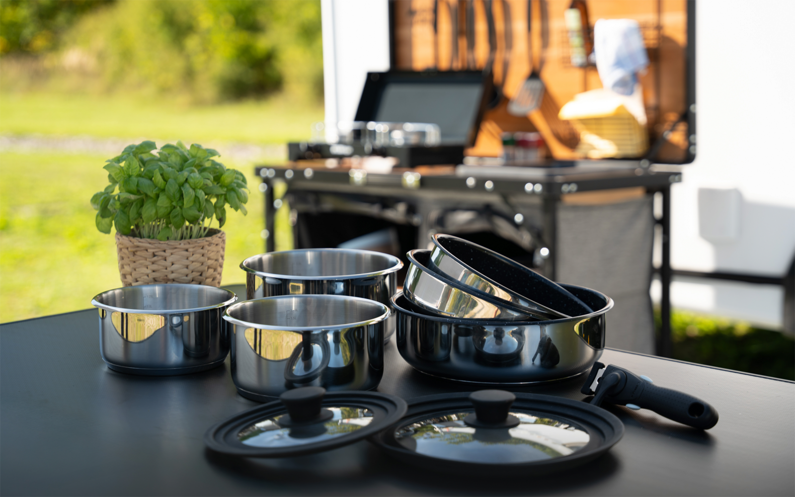 Mehrere Edelstahltöpfe und Pfannen mit Glasdeckeln und Griffen auf einem dunklen Tisch; links ein kleiner Basilikumtopf; im Hintergrund ein offener Stauraum eines Wohnmobils und grüner Außenbereich