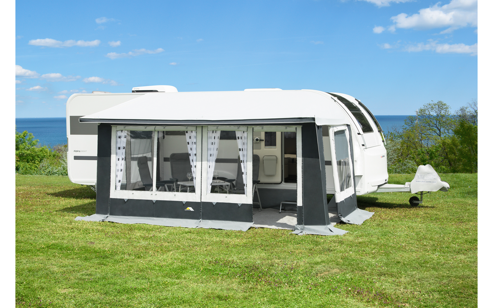 Weißer Wohnwagen mit angebautem Vorzelt auf einer Rasenfläche, im Vorzelt sichtbare Stühle, im Hintergrund Meer, Bäume und blauer Himmel mit Wolken