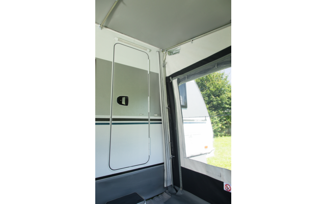 Innenseite eines Vorzelts mit Blick auf die geschlossene Tür eines weißen Wohnwagens, Griff, horizontale Streifen, Metallstützen, aufgerolltes Seitenteil und Gras mit Bäumen draußen