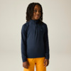 Kind met halflange dreadlocks, draagt een donkerblauw langermig shirt met halve ritssluiting en klein borstlogo en mosterdgele broek, staat voor een lichte achtergrond, handen langs het lichaam, kijkt iets naar rechts