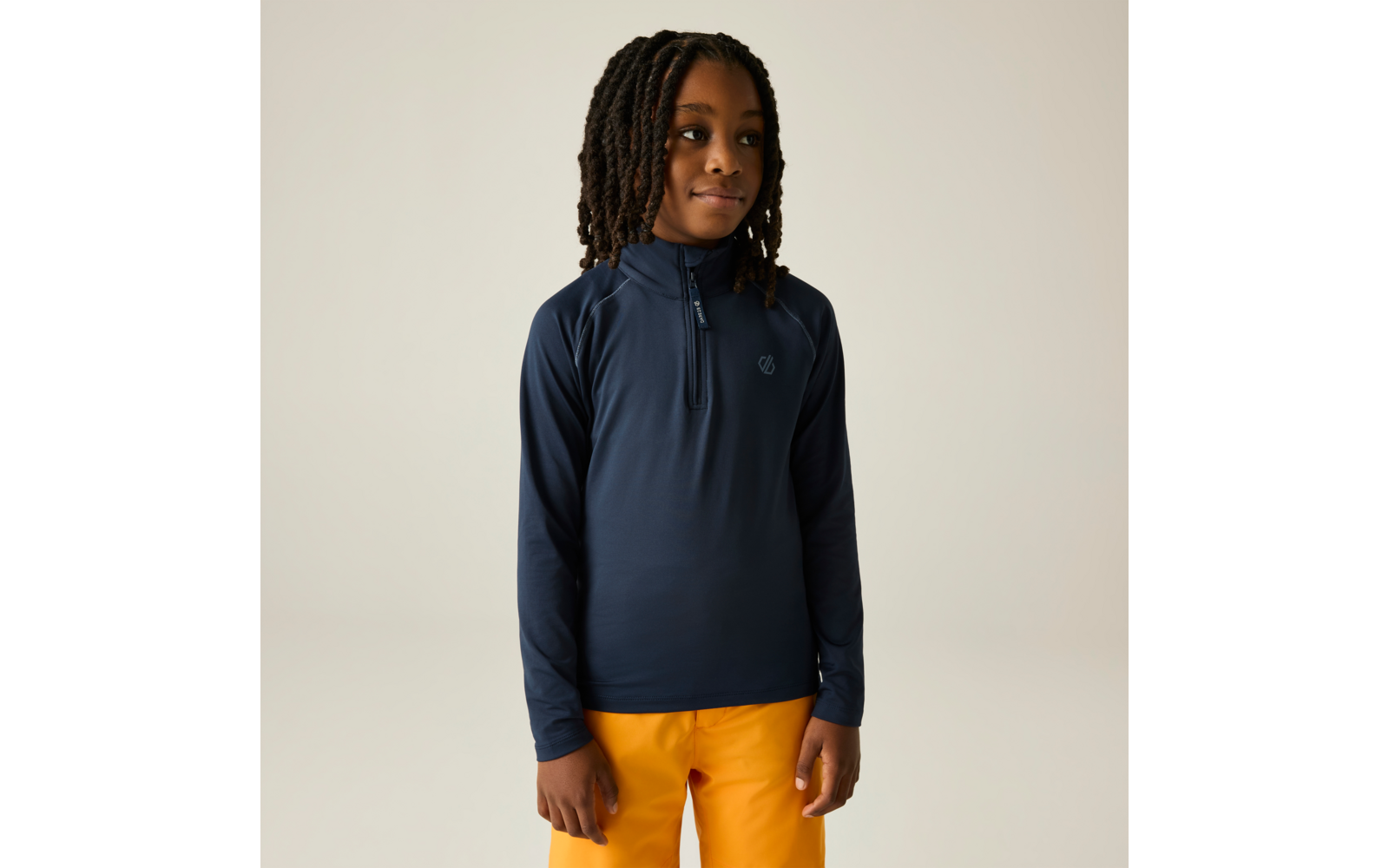 Kind met halflange dreadlocks, draagt een donkerblauw langermig shirt met halve ritssluiting en klein borstlogo en mosterdgele broek, staat voor een lichte achtergrond, handen langs het lichaam, kijkt iets naar rechts