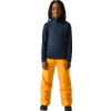 Persoon staat recht voor een lichte achtergrond, draagt een donkerblauw shirt met rits en lange mouwen, feloranje broek, zwarte schoenen, haar in vlechten