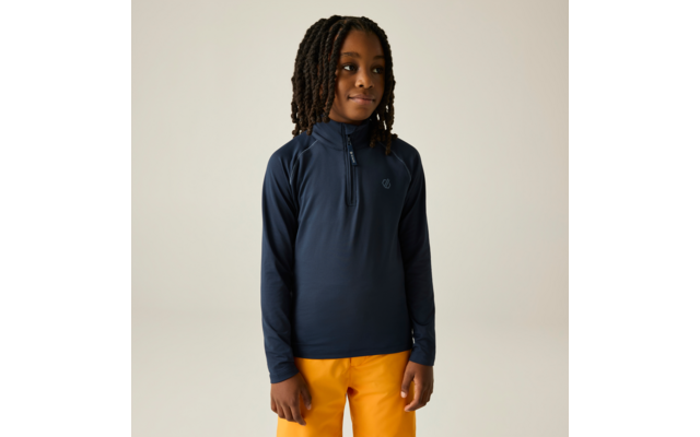 Kind met halflange dreadlocks, draagt een donkerblauw langermig shirt met halve ritssluiting en klein borstlogo en mosterdgele broek, staat voor een lichte achtergrond, handen langs het lichaam, kijkt iets naar rechts