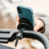 Hand hält Smartphone mit blaugrüner, gesprenkelter Hülle, an einem runden schwarzen Halter am Fahrradlenker befestigt