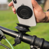 Hand hält weißes Smartphone mit schwarzem kreisförmigem Magnetpad auf der Rückseite über einer schwarzen Fahrradlenkerhalterung; grünes Bremskabel und unscharfer Gras-Hintergrund