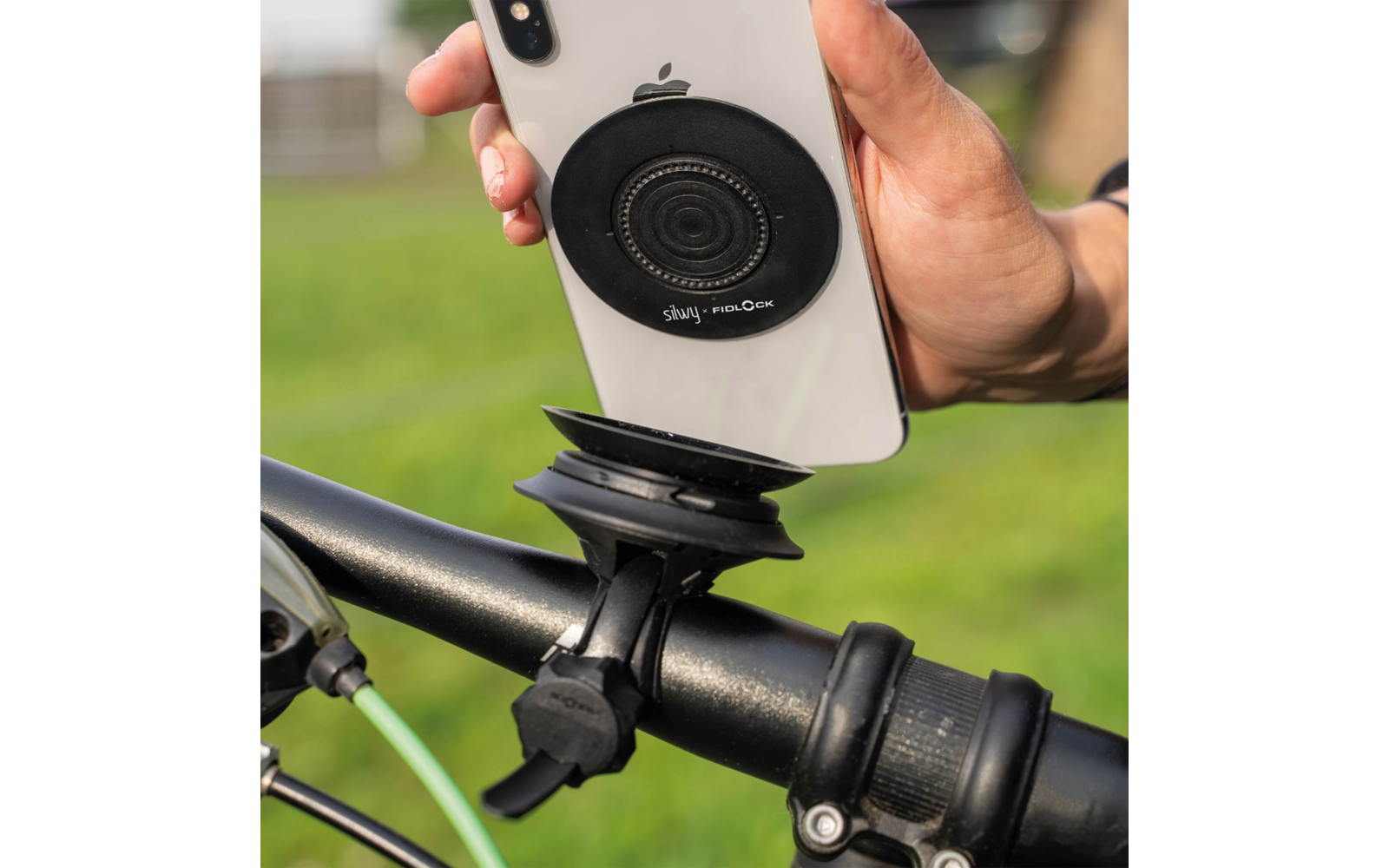 Hand hält weißes Smartphone mit schwarzem kreisförmigem Magnetpad auf der Rückseite über einer schwarzen Fahrradlenkerhalterung; grünes Bremskabel und unscharfer Gras-Hintergrund