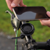 Zwei Hände halten ein Smartphone über einer runden Handyhalterung am Fahrradlenker, im Hintergrund Gras