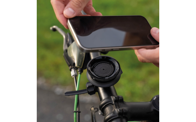 Zwei Hände halten ein Smartphone über einer runden Handyhalterung am Fahrradlenker, im Hintergrund Gras