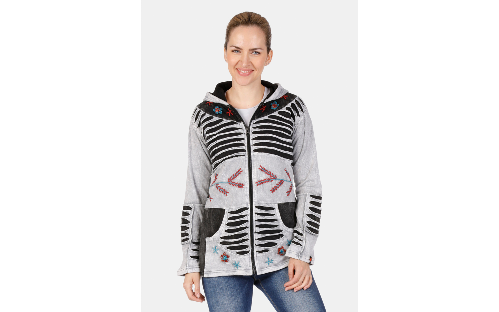 Frau vor weißem Hintergrund, trägt graue Jacke mit Reißverschluss, schwarzem Streifenmuster, bunten Stickereien und blaue Jeans