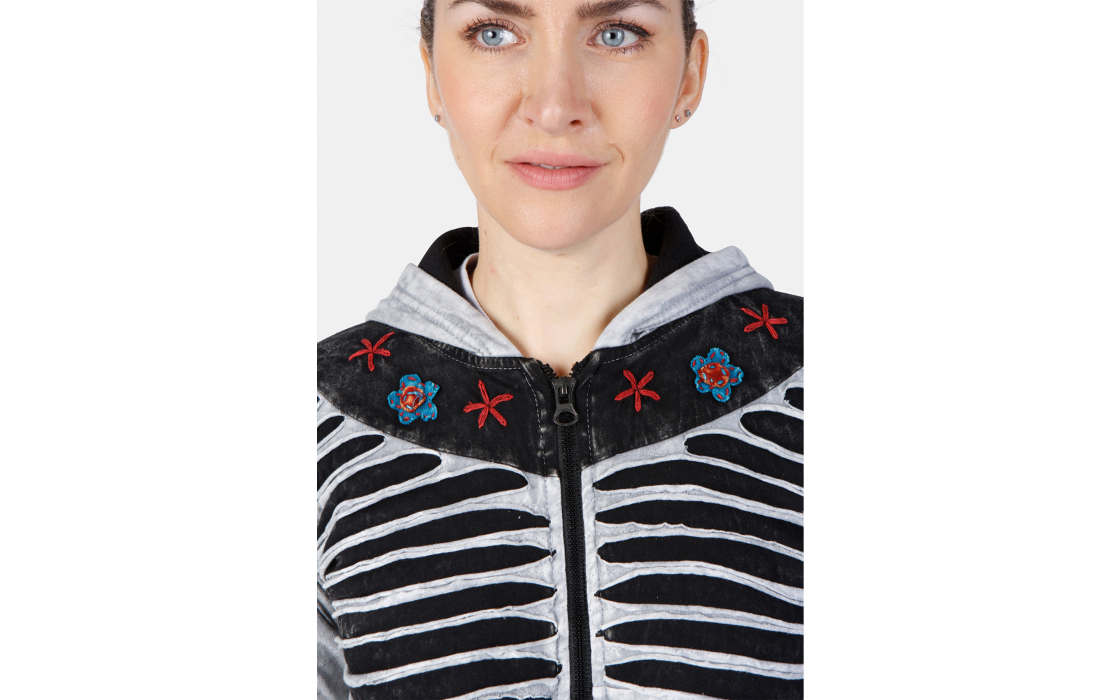 Nahaufnahme einer Person von Kinn bis Stirn mit sichtbaren blauen Augen, Nase und Mund. Trägt eine schwarze Reißverschlussjacke mit weißen gebogenen Streifen, roten Blumenstickereien und hellgrauer Kapuze