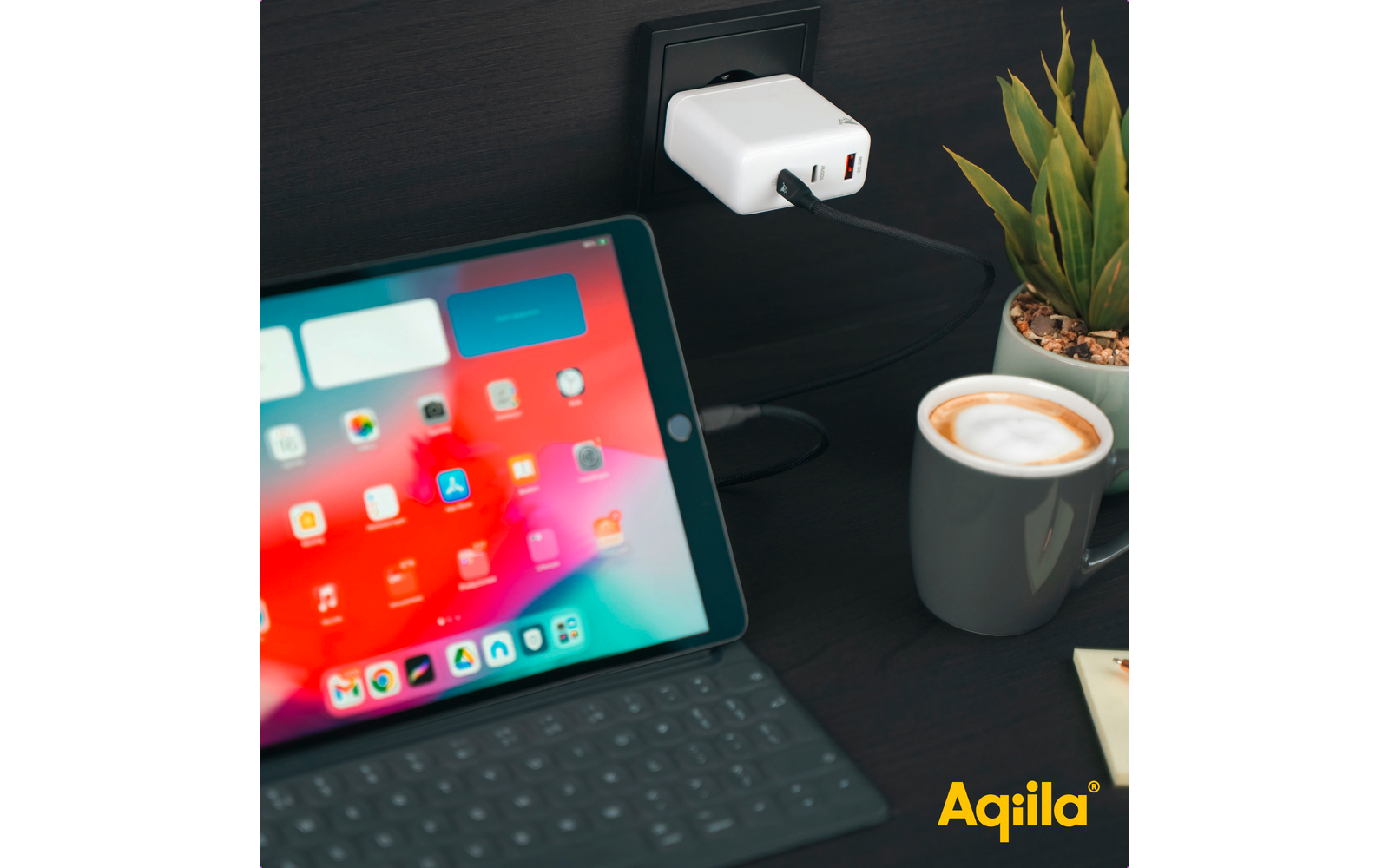 Tablet mit geöffnetem Bildschirm und Tastatur, Ladekabel angeschlossen, neben einer Tasse Kaffee und einer Pflanze