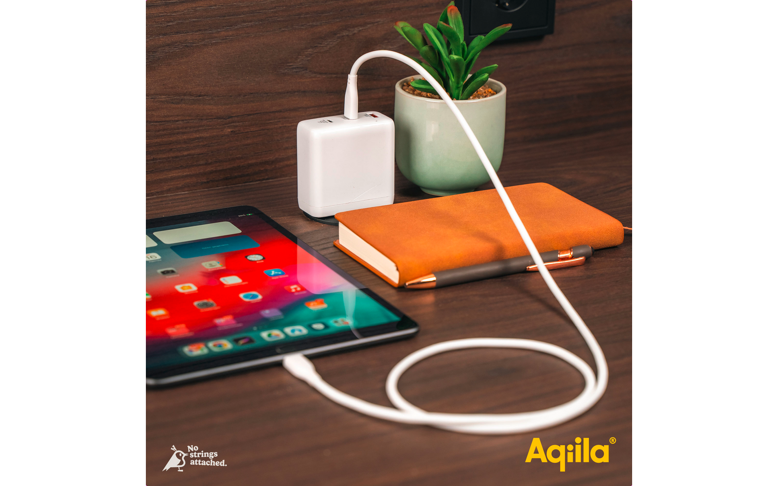 Smartphone mit Apps auf dem Bildschirm, daneben ein orangefarbenes Notizbuch, eine Pflanze in einem weißen Topf und ein weißes Ladegerät mit Kabel