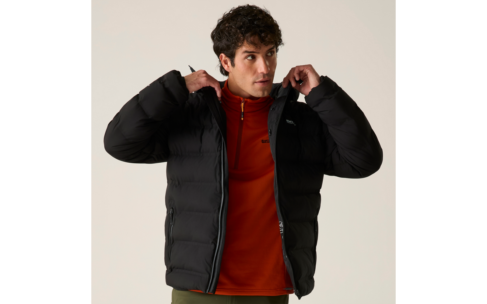 Mann trägt schwarze Steppjacke, teilweise geöffnet, darunter orangefarbene Fleecejacke, hält den Kragen, schaut zur Seite, heller Hintergrund