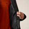 Hand hält die Innenseite einer schwarzen Jacke offen; darunter ein orangefarbener Pullover; Innenfutter mit Reißverschlusstasche und drei runden Symbolen
