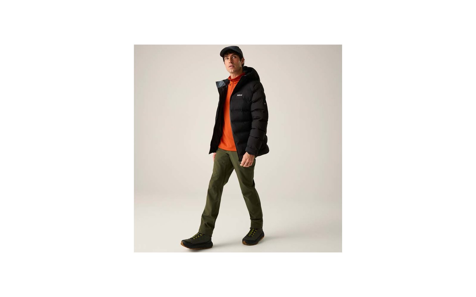 Person in schwarzer Steppjacke, orangefarbenem Pullover, olivgrünen Hosen, schwarzen Schuhen und schwarzer Schirmmütze vor hellem Hintergrund, gehend
