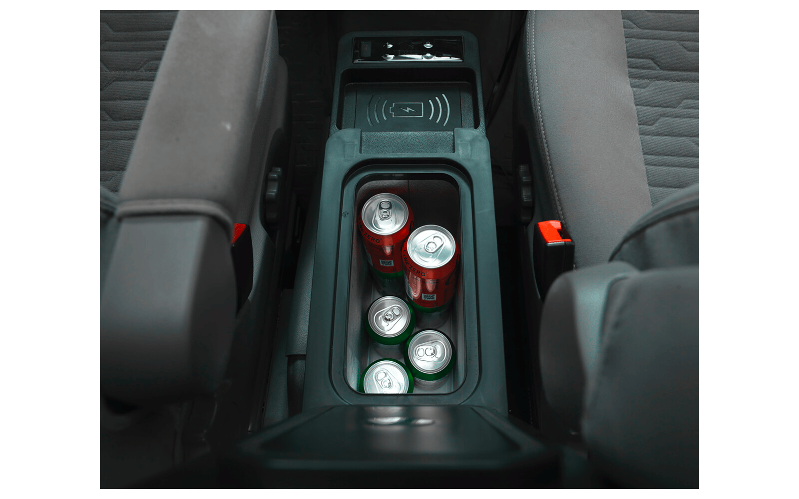 Mittelkonsole eines Autos zwischen den Vordersitzen mit vier Getränkedosen in den Haltern: zwei rote Dosen und zwei silberne Dosen mit grünen Oberseiten; sichtbare Sitzbezüge und rote Gurtverschlüsse