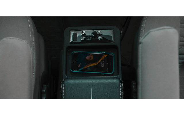 Mittelkonsole eines Autos zwischen zwei grauen Vordersitzen; ein Smartphone liegt in einem Ablagefach, Mittelarmlehne und Bedienfeld oberhalb sichtbar