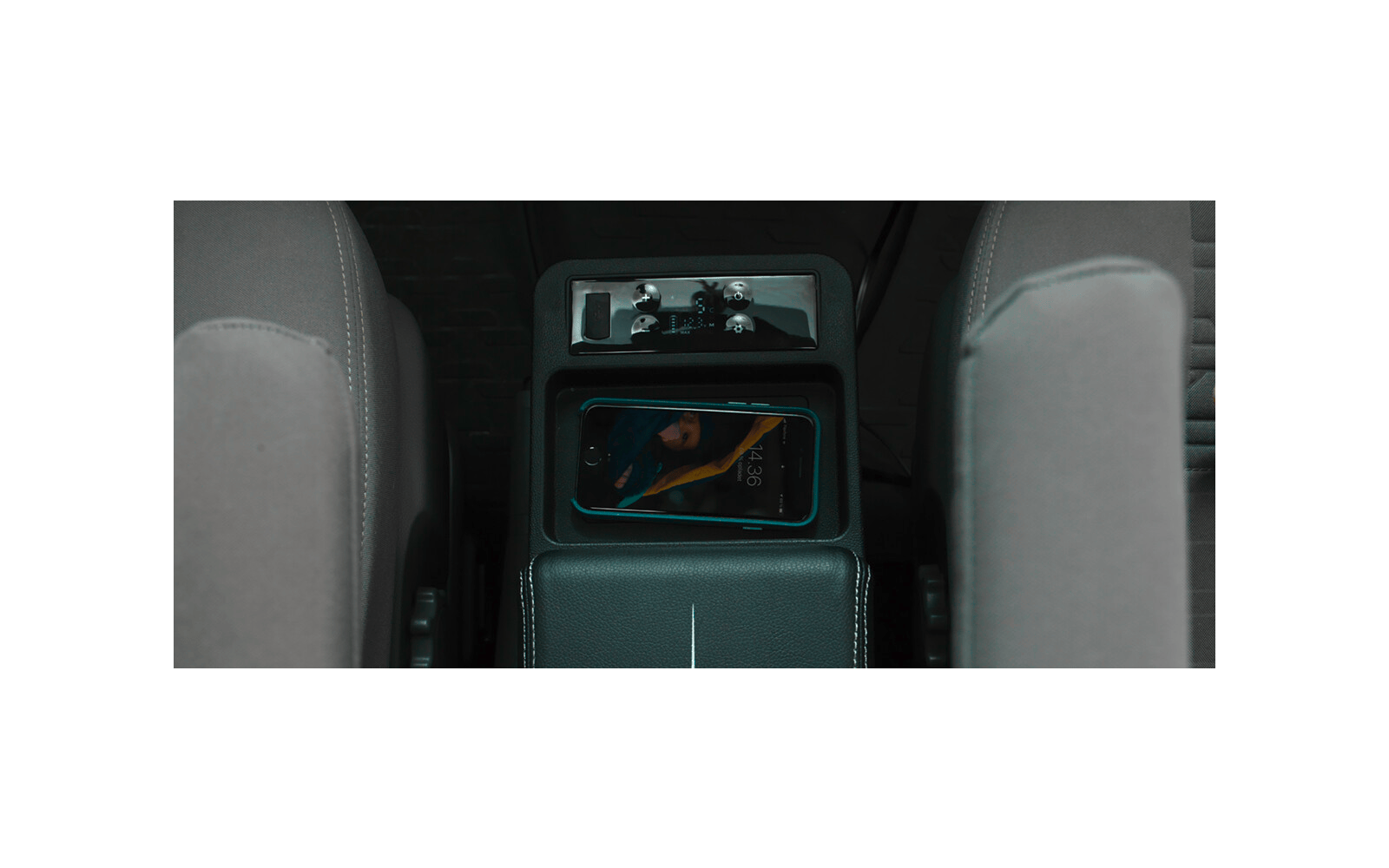 Mittelkonsole eines Autos zwischen zwei grauen Vordersitzen; ein Smartphone liegt in einem Ablagefach, Mittelarmlehne und Bedienfeld oberhalb sichtbar