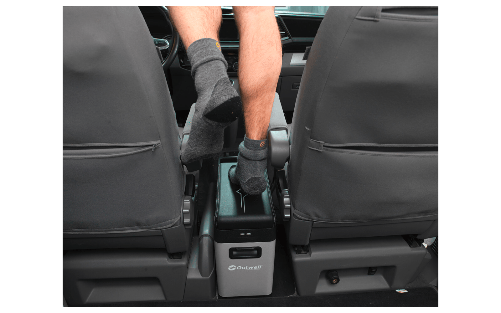 Unterschenkel und Füße einer sitzenden Person mit schwarzen Socken auf der Mittelkonsole eines Autos; zwei Vordersitze mit grauen Bezügen und eine graue Outwell-Kühlbox zwischen den Sitzen