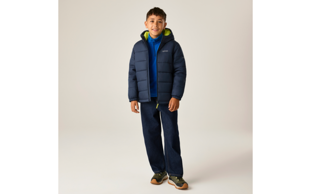 Vollkörperaufnahme eines Jungen in dunkelblauer Steppjacke mit hellgrünem Innenfutter, blauer Kapuzenjacke darunter, dunklen Jeans und grünen Sportschuhen vor hellem Hintergrund