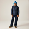 Eine stehende Person, lächelnd, trägt eine dunkelblaue Steppjacke, eine hellblaue Strickmütze mit Bommel, dunkelblaue Hose und grüne Sneaker vor hellem Hintergrund