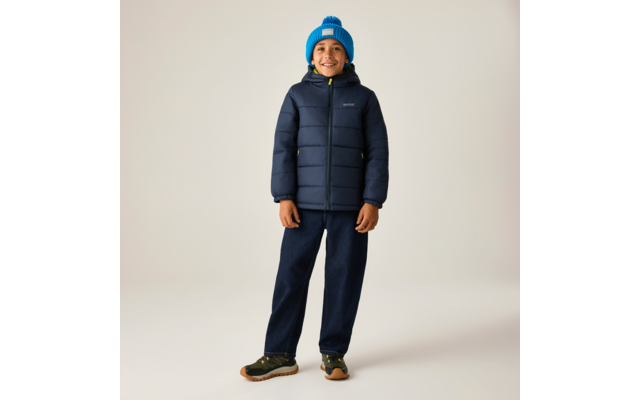 Eine stehende Person, lächelnd, trägt eine dunkelblaue Steppjacke, eine hellblaue Strickmütze mit Bommel, dunkelblaue Hose und grüne Sneaker vor hellem Hintergrund