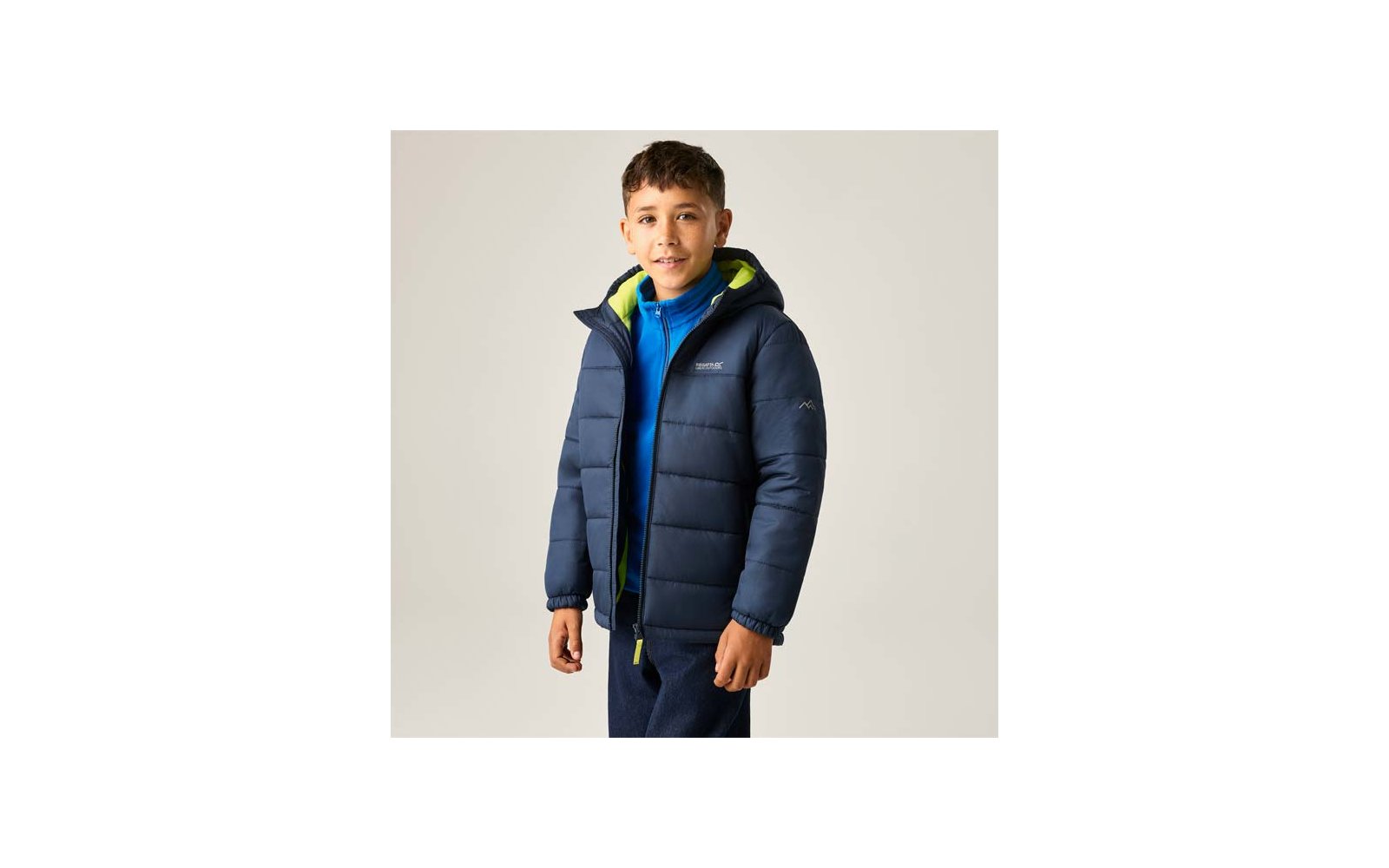 Person mit kurzen dunklen Haaren, trägt eine marineblaue gesteppte Jacke mit grün gefütterter Kapuze und einen blauen Reißverschluss-Pullover, steht vor hellem Hintergrund