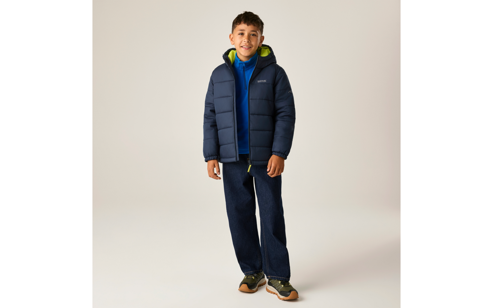 Vollkörperaufnahme eines Jungen in dunkelblauer Steppjacke mit hellgrünem Innenfutter, blauer Kapuzenjacke darunter, dunklen Jeans und grünen Sportschuhen vor hellem Hintergrund