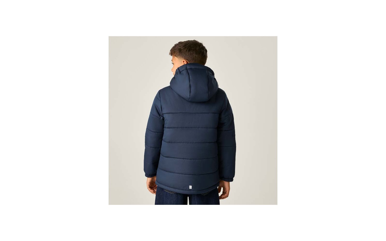 Person von hinten, trägt dunkelblaue gesteppte Jacke mit Kapuze, kleines weißes Etikett am unteren Rücken, heller einfarbiger Hintergrund