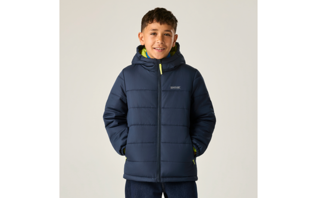 Junge vor hellem Hintergrund, trägt dunkelblaue gesteppte Kapuzenjacke mit grüner Innenkante, Hände in den Taschen, lächelnd