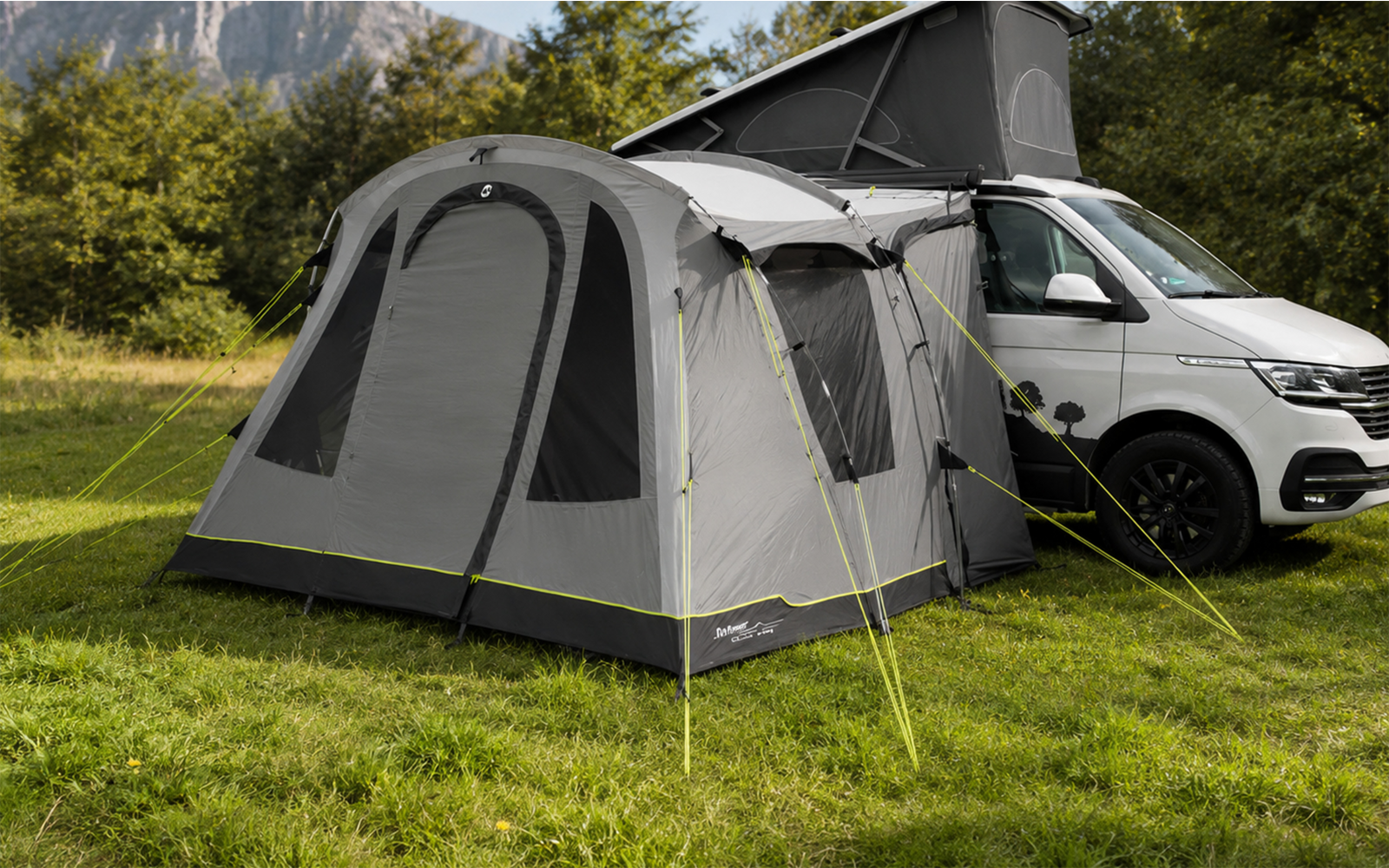 Graues Vorzelt an weißem Campingbus mit aufgeklapptem Dach, auf einer Wiese vor Bäumen und Bergen