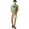 Mann steht vor hellem Hintergrund, trägt hellgrünes T‑Shirt mit dunklem rechteckigen Aufdruck, beige Shorts, schwarze Cap, graue Socken und dunkle Sneaker, Hände vor der Taille, Blick zur Seite
