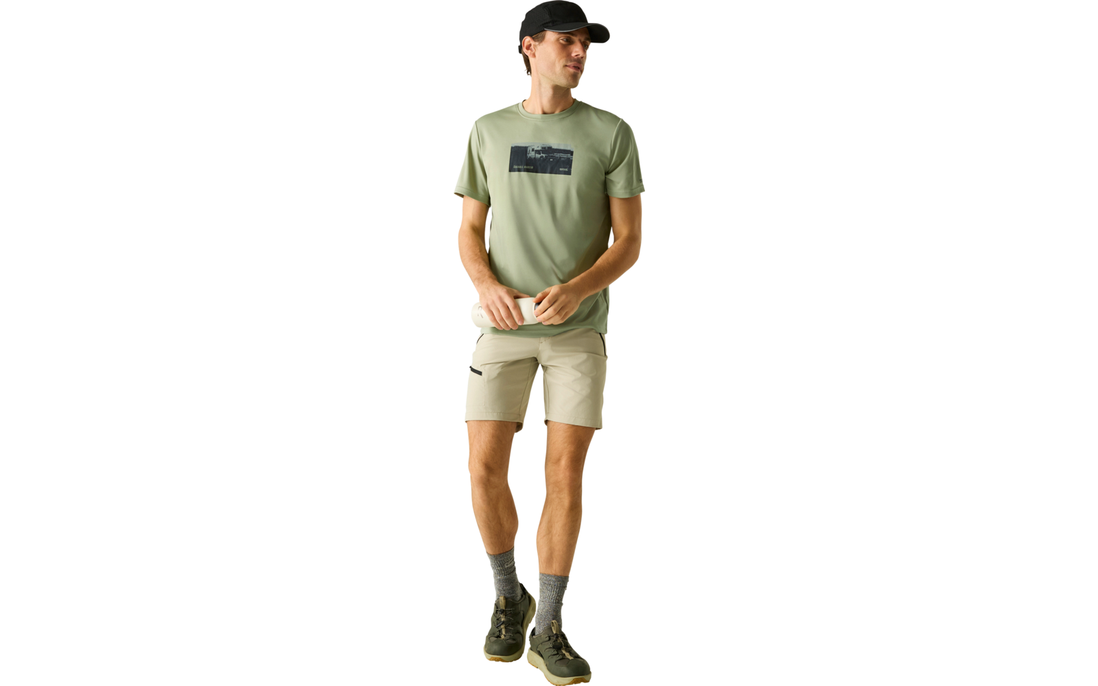 Mann steht vor hellem Hintergrund, trägt hellgrünes T‑Shirt mit dunklem rechteckigen Aufdruck, beige Shorts, schwarze Cap, graue Socken und dunkle Sneaker, Hände vor der Taille, Blick zur Seite