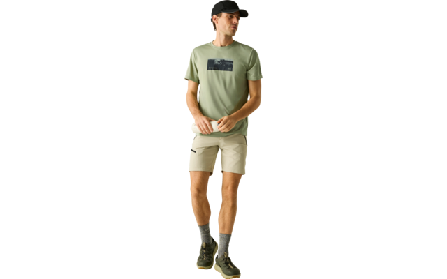 Mann steht vor hellem Hintergrund, trägt hellgrünes T‑Shirt mit dunklem rechteckigen Aufdruck, beige Shorts, schwarze Cap, graue Socken und dunkle Sneaker, Hände vor der Taille, Blick zur Seite