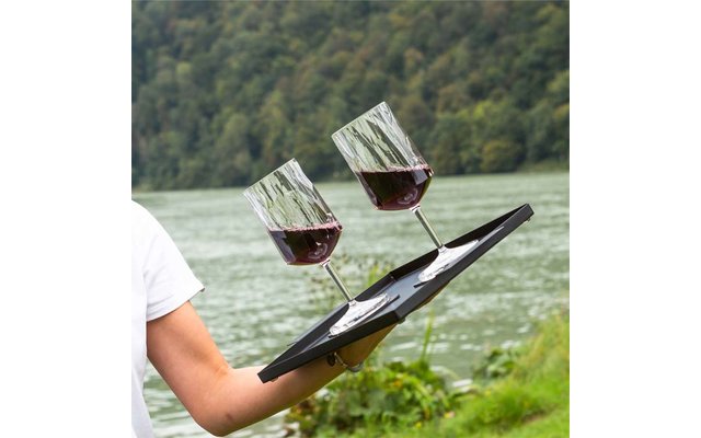 Arm of a person in a white shirt holding a black tray with two wine glasses partially filled with red wine in front of a lake and tree-covered shore