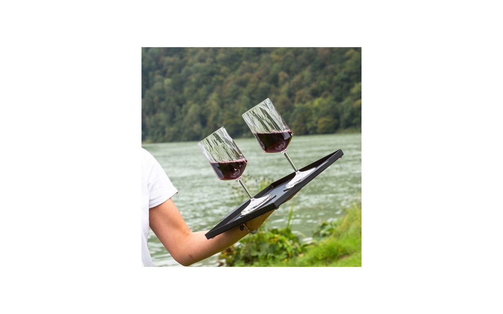 Arm of a person in a white shirt holding a black tray with two wine glasses partially filled with red wine in front of a lake and tree-covered shore