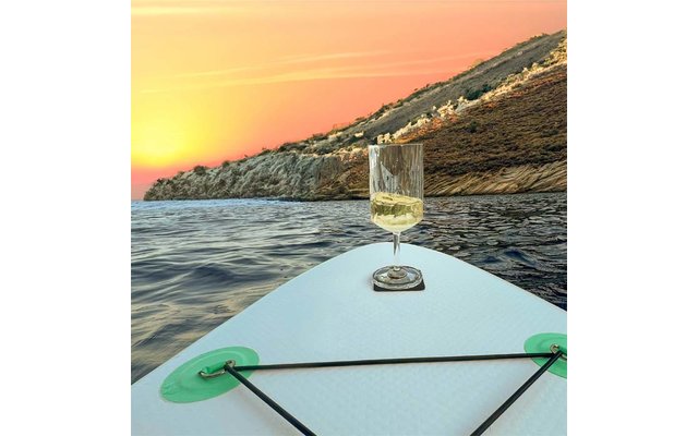 Front of a stand-up paddleboard with a glass containing a pale drink, open sea, rocky coastline, sunset