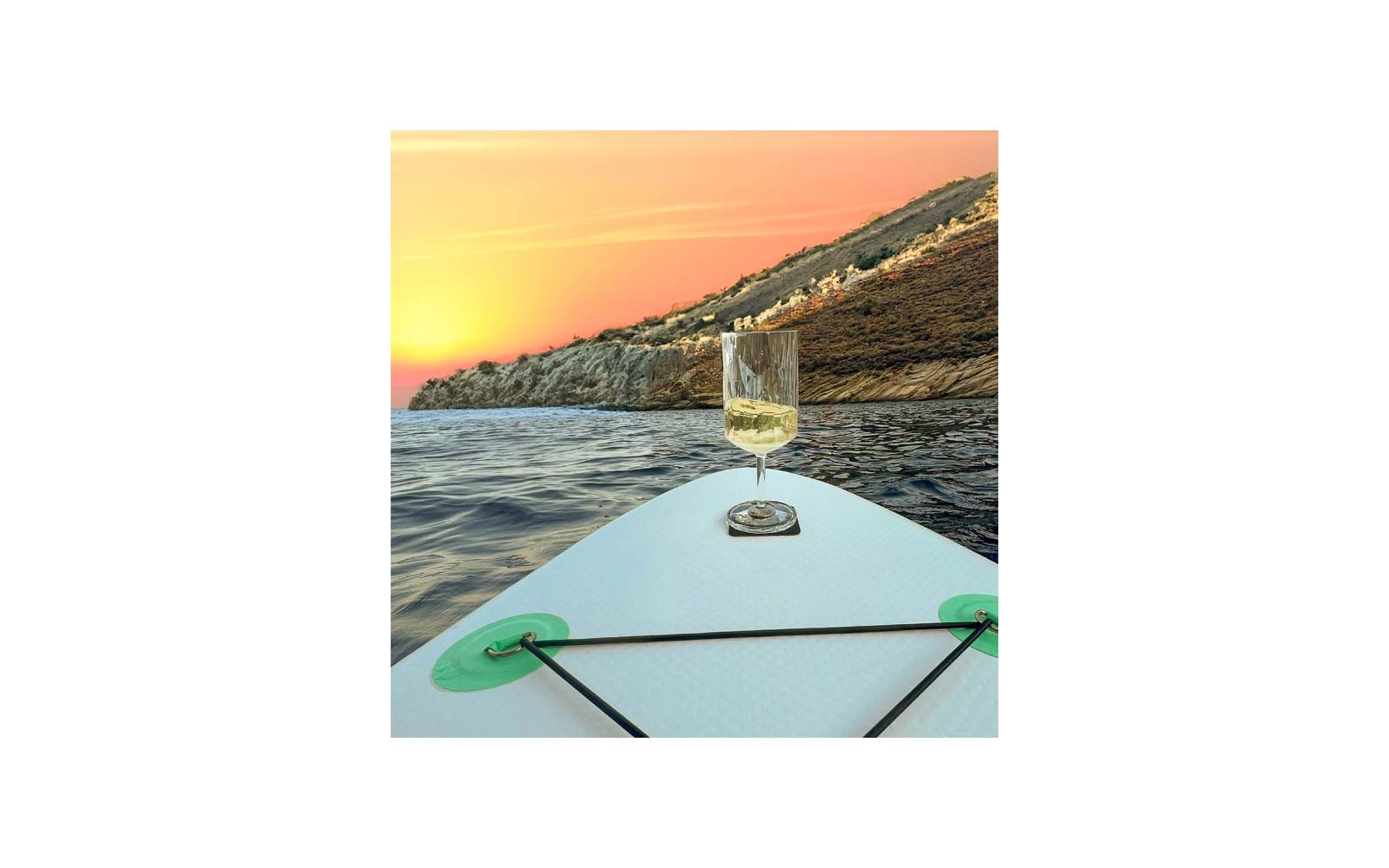 Front of a stand-up paddleboard with a glass containing a pale drink, open sea, rocky coastline, sunset